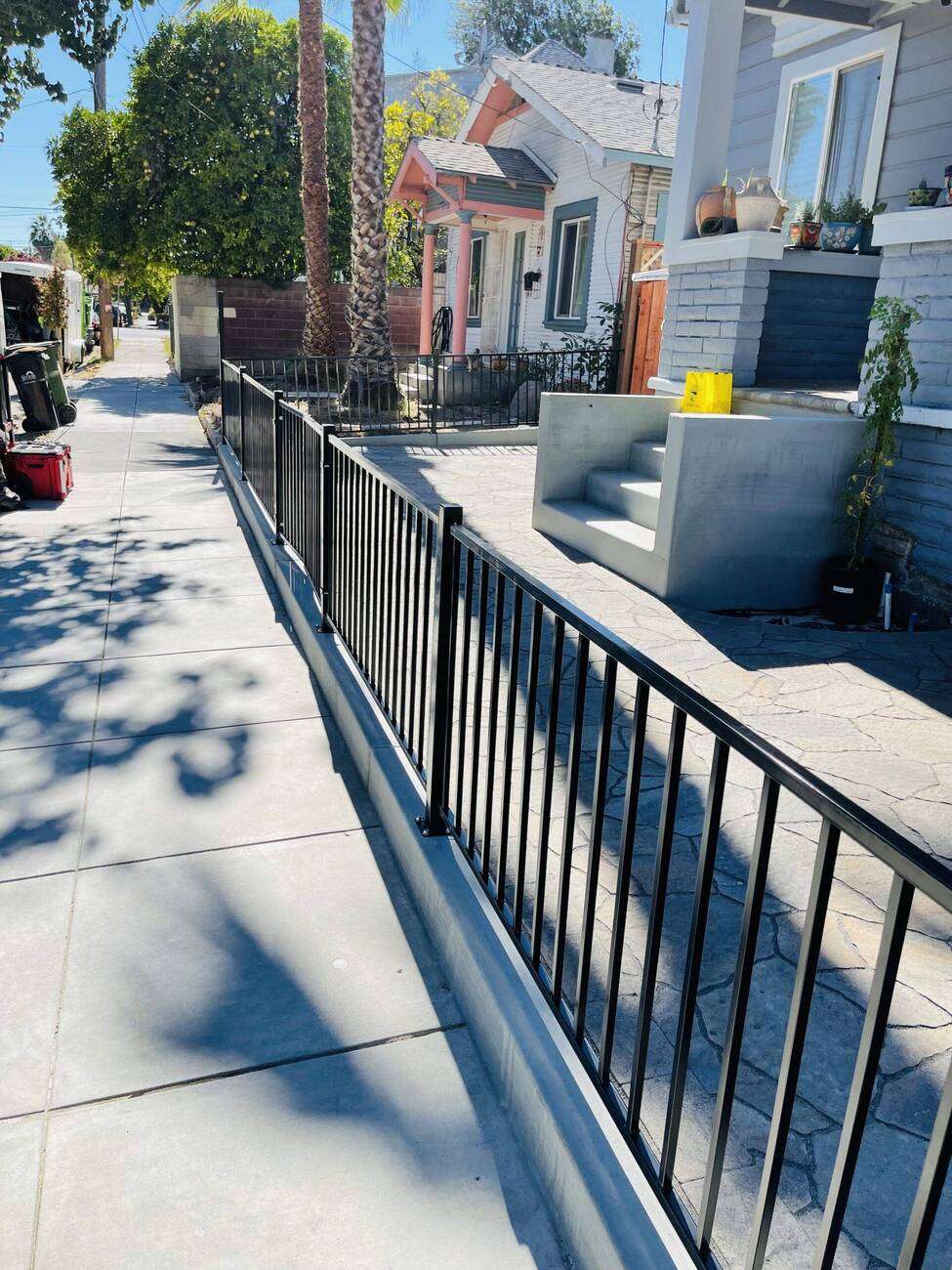 Black metal fence along a concrete pathway leading to a residential home, with palm trees and a landscaped front yard, emphasizing outdoor space design and maintenance.