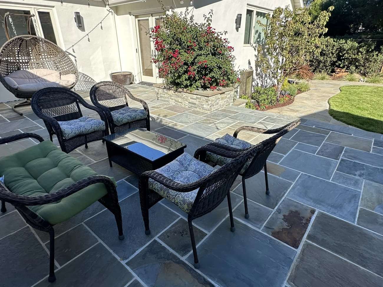 Outdoor patio featuring comfortable wicker seating with patterned cushions, a green cushioned chair, a glass-top coffee table, and surrounding lush landscaping, illustrating a stable and inviting outdoor space ideal for gardening and erosion control.