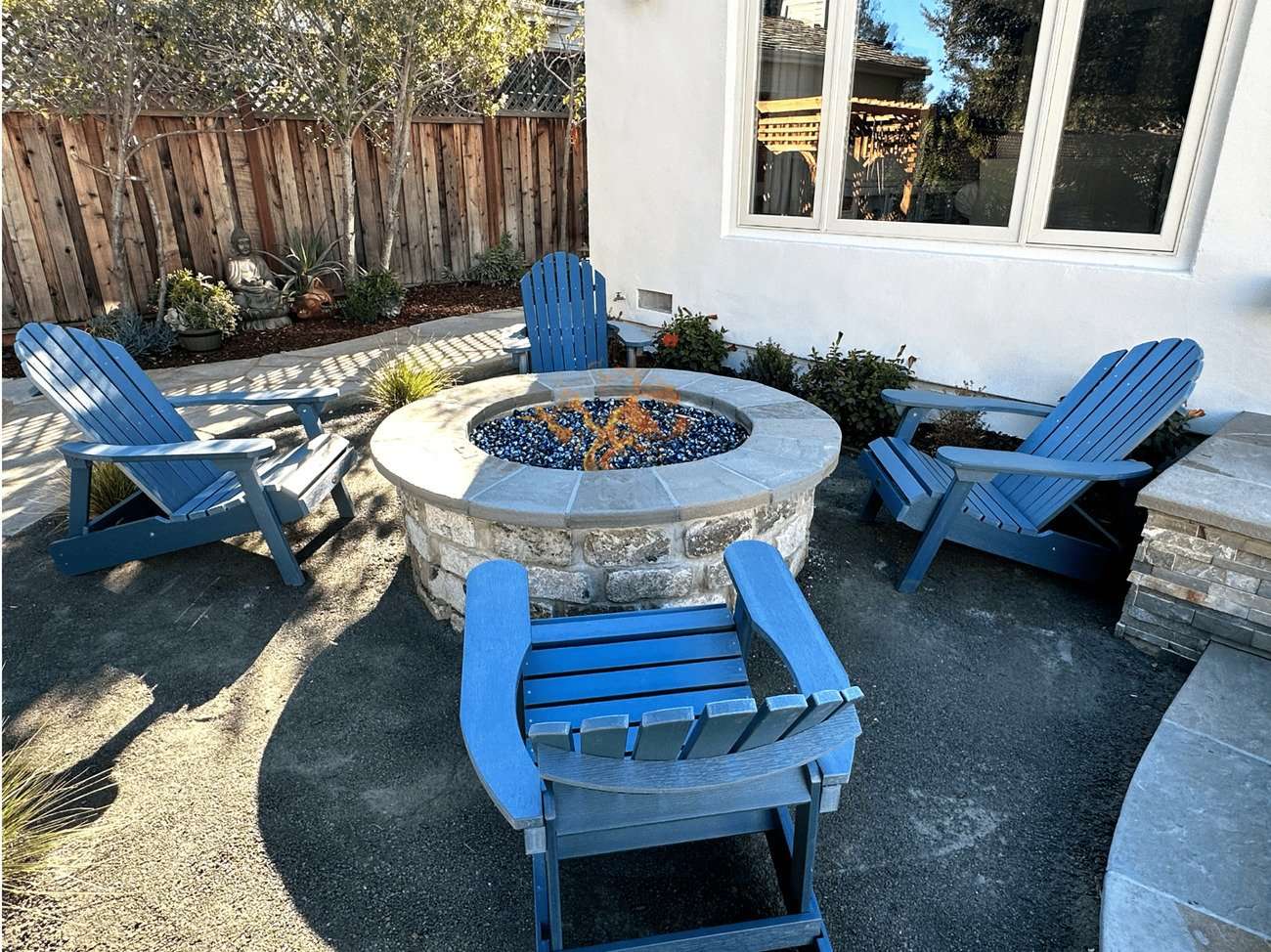 Blue Adirondack chairs surrounding a stone fire pit with decorative glass stones, set in a landscaped outdoor space with plants and a wooden fence in the background.
